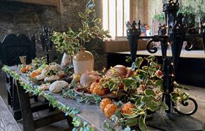 A long wooden table laden with a festive spread of bread, oranges and a roasted fowl, decorated with holly branches and fairy lights.